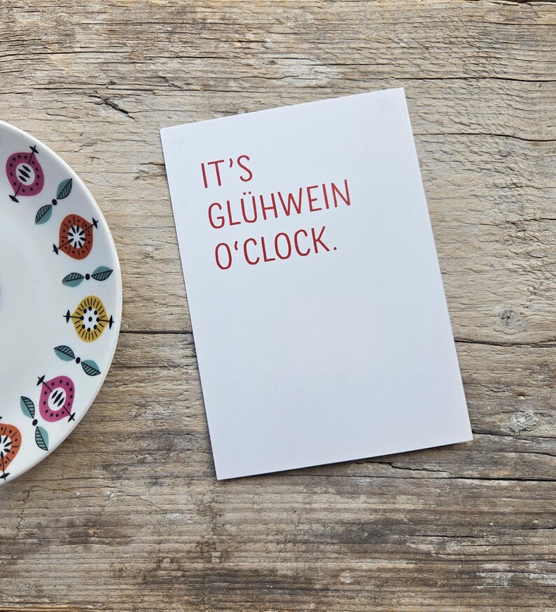 May include: A white rectangular greeting card with the red text IT'S GL&Uuml;HWEIN O'CLOCK. positioned next to a white ceramic plate featuring a repeating pattern of stylized pink, orange, and yellow circular shapes with small green leaves.