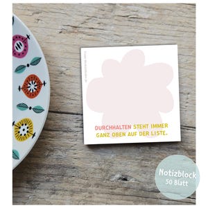 May include: A square notepad with a light pink floral design and the text "DURCHHALTEN STEHT IMMER GANZ OBEN AUF DER LISTE." A decorative plate with colorful floral designs is in the background. The notepad has 50 sheets.