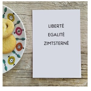 May include: A white rectangular card with the black printed text LIBERT&Eacute; EGALIT&Eacute; ZIMTSTERNE positioned next to a white ceramic plate featuring a colorful floral pattern and two round, light-colored cookies with a textured surface.