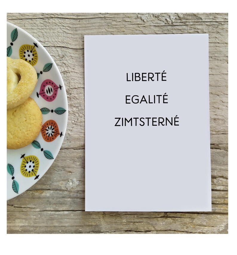 May include: A white rectangular card with the black printed text LIBERT&Eacute; EGALIT&Eacute; ZIMTSTERNE positioned next to a white ceramic plate featuring a colorful floral pattern and two round, light-colored cookies with a textured surface.