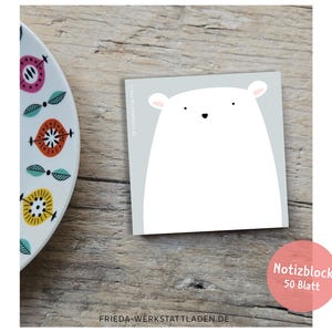 May include: A square notepad featuring a white bear illustration on a light grey backdrop. The notepad rests on a wooden surface beside a decorative plate with colourful floral patterns. The notepad has the text "Notizblock 50 Blatt".