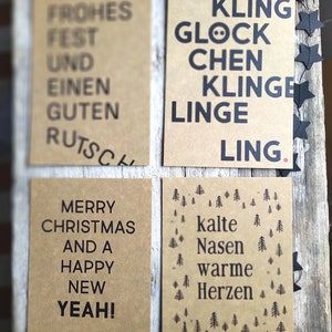 May include: Four brown cards with black text. The first card says "Frohes Fest und einen guten Rutsch". The second card says "Kling Glock Chen Klinge Linge Ling". The third card says "Merry Christmas and a Happy New Yeah!". The fourth card says "Kalte Nasen Warme Herzen" and has a pattern of small pine trees.