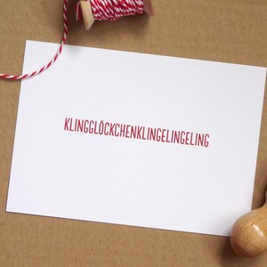 May include: A rectangular white card featuring the red text KLINGGL&Ouml;CKCHENKLINGELINGELING centered on the surface. A wooden rubber stamp sits in the bottom right corner, and a spool of red and white baker's twine is positioned at the top.