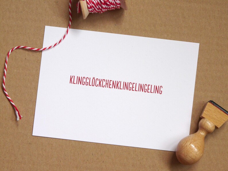 May include: A rectangular white card featuring the red text KLINGGL&Ouml;CKCHENKLINGELINGELING centered on the surface. A wooden rubber stamp sits in the bottom right corner, and a spool of red and white baker's twine is positioned at the top.