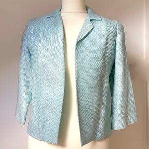 Könnte beinhalten: Ein hellblaues Blazer mit strukturiertem Gewebe. Die Jacke mit offenem Schnitt hat einen Reverskragen und Dreiviertelärmel. Der Blazer wird auf einer Schaufensterpuppe präsentiert.