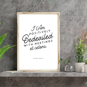 以下が含まれることがあります： 「I Am Positively Bedeviled With Meetings et cetera. Moira Rose.」のテキストが入った額入りのプリント。プリントは明るい木製のフレームに入っており、植物や照明器具のある灰色の棚に飾られています。