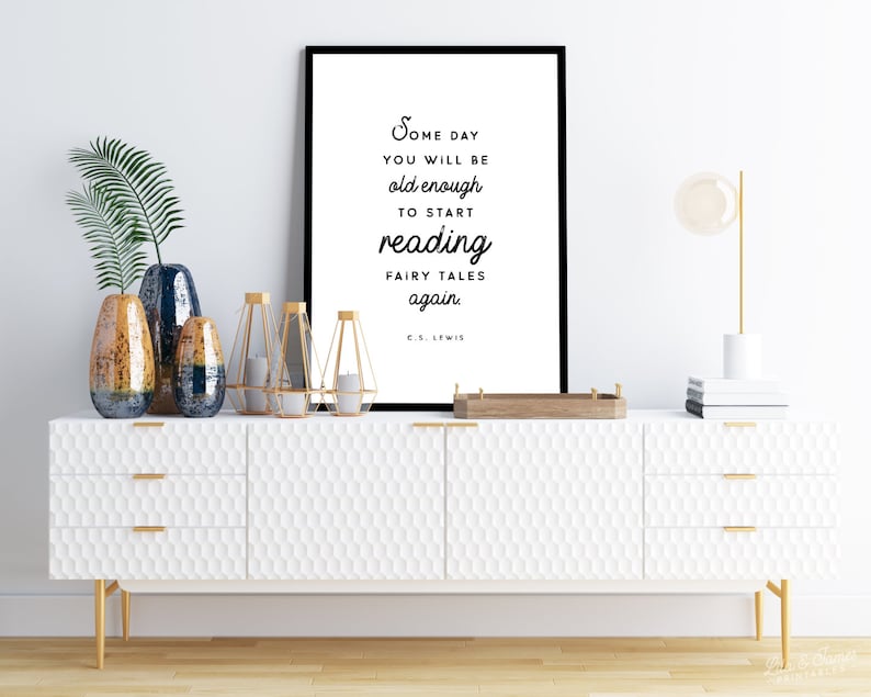 May include: A white framed print with a black and white quote by C.S. Lewis that reads "Some day you will be old enough to start reading fairy tales again." The print is hanging on a white wall above a white dresser with gold accents.