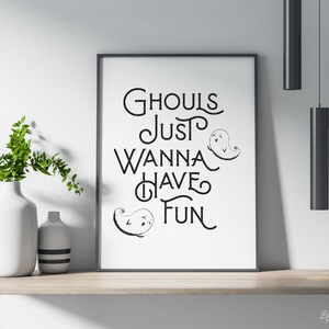 May include: A framed print with the text "Ghouls Just Wanna Have Fun" in black script, accompanied by ghost illustrations. The print is displayed on a wooden shelf with white vases and greenery, creating a Halloween theme.