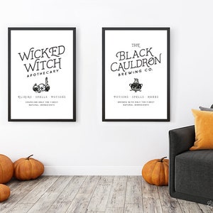 May include: Two black framed prints with white backgrounds and black text. The print on the left says "Wicked Witch Apothecary" with a skull and potion bottles. The print on the right says "The Black Cauldron Brewing Co." with a cauldron and a witch's hat. The prints are hanging on a white wall with a gray couch and orange throw pillows in front of them.