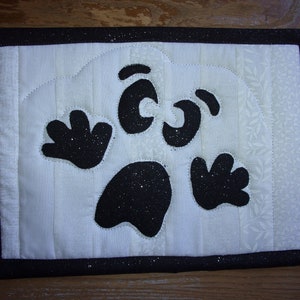 May include: A white quilted fabric ghost with black glitter accents. The ghost has two eyes, a mouth, and two hands. The ghost is framed with a black border.