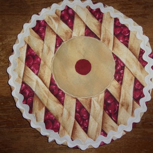 May include: A round, quilted fabric trivet designed to resemble a cherry pie. The trivet features a lattice crust pattern in tan and brown, with visible red cherries. A beige center with a red dot is surrounded by a white, scalloped edge.