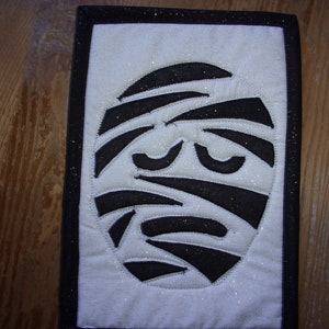 May include: A white fabric square with a black mummy face design. The mummy face has a black outline and is covered in white bandages. The fabric has a glittery texture.