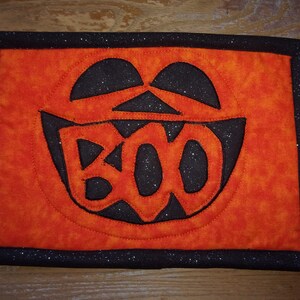 May include: A rectangular orange and black Halloween-themed quilted placemat. The design features a jack-o'-lantern face with the word "BOO" in the mouth. The border is black with glitter.