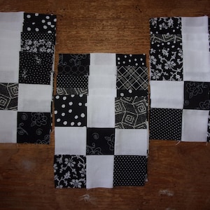 May include: Three fabric squares in a checkerboard pattern, featuring black and white squares. The black squares have various patterns, including polka dots, floral designs, and geometric shapes. The squares are arranged on a wooden surface.