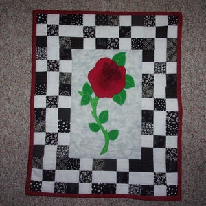 May include: A handmade quilt featuring a large red rose with green leaves and stem. The rose is centered on a light gray background, framed by a checkerboard pattern of black and white squares. The quilt has a red border.