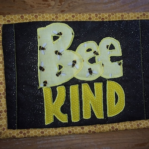 May include: A black and yellow fabric coaster with the words "Bee Kind" embroidered on it. The letters are outlined in yellow and the background is a black fabric with a subtle sparkle.