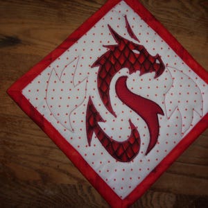 May include: A diamond-shaped quilted potholder featuring a white fabric with small red polka dots. The center displays an embroidered red dragon silhouette with a scale pattern, flanked by two stitched flame shapes, all bordered by red fabric.