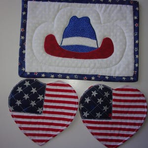 May include: A patriotic-themed set featuring a quilted placemat with a cowboy hat design in red, white, and blue, and two heart-shaped coasters with an American flag pattern. The placemat has a blue border with white stars.