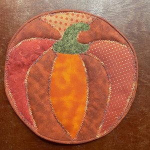 May include: A circular fabric coaster or mat featuring a quilted pumpkin design. The pumpkin is constructed from various orange, red, and brown fabric patches with visible zigzag stitching. A green fabric stem sits at the top center.