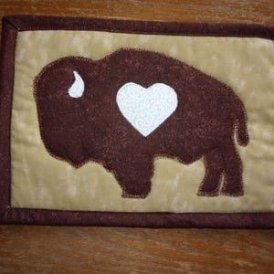 May include: A brown felt coaster with a brown buffalo silhouette and a white heart in the center. The coaster has a tan background and a brown border.