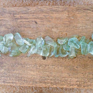 Puede incluir: Una tabla de madera rectangular con un patrón de vetas naturales. Una línea de piezas de vidrio verde claro de forma irregular está dispuesta a lo largo del centro de la tabla, creando un elemento decorativo. Las piezas de vidrio son translúcidas.