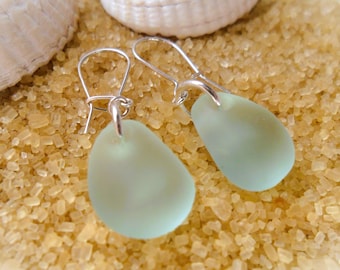 May include: A pair of seafoam green teardrop-shaped earrings with silver-colored hooks. The earrings are displayed on a bed of sand with seashells in the background. The earrings have a smooth, frosted appearance.