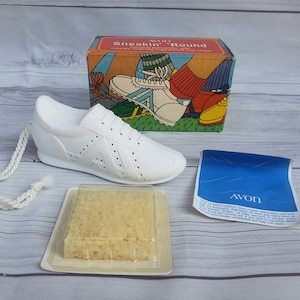 May include: White sneaker-shaped soap with a white string attached. The soap is in a clear plastic container with a white label. The label says "Avon Sneak'n' Round" and shows a cartoon of a person wearing sneakers. The soap is also in a blue and white box with the word "Avon" on it.
