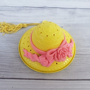 May include: A yellow plastic hat with a pink ribbon and flower decoration. The hat has a straw-like texture and holes throughout.