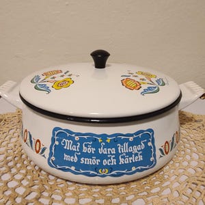 May include: White enamel cooking pot with a black rim and lid, featuring floral designs in yellow, blue, and orange. The pot has side handles and a black knob. A blue label with white text is on the front. The pot is on a beige doily.