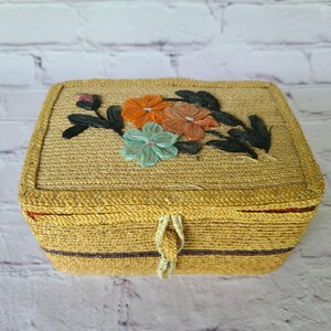 May include: A rectangular woven straw box with a hinged lid featuring embroidered floral details in orange, light brown, and teal with dark green leaves. The box has a natural tan color with a small loop closure and a thin dark brown stripe.