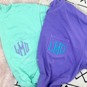 May include: Two t-shirts with a pocket on the front. The t-shirt on the left is light green with a purple monogram that says "LMD". The t-shirt on the right is purple with a turquoise monogram that says "LMD".