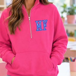 May include: A bright hot pink quarter-zip sweatshirt featuring a silver-toned metal zipper and a front kangaroo pocket. The left chest area displays the embroidered letters MF in a bold blue font with a light blue outline on the fabric.