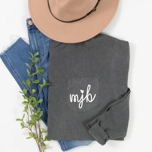 May include: A charcoal gray pocket t-shirt with the initials "mjb" in white script, topped with a tan hat and blue jeans. A sprig of greenery is placed near the jeans. The shirt is folded, showing the pocket detail.
