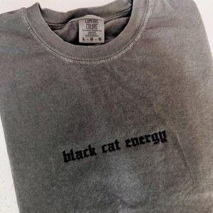 May include: A gray Comfort Colors long-sleeve shirt with the text "black cat energy" in a gothic font. The shirt has a crew neck and a tag at the neckline indicating size L-G-G.