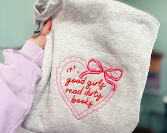 May include: A light gray sweatshirt with a pink embroidered heart design. The heart features the phrase "good girls read dirty books" in red script, with a pink bow and decorative stars. The sweatshirt is a Gildan brand.