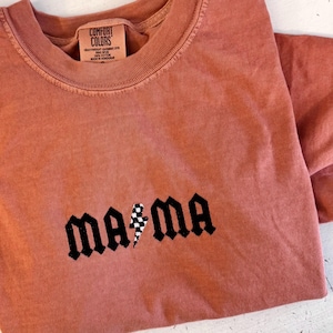 May include: A terracotta-colored Comfort Colors t-shirt with the word "mama" in a gothic font, featuring a checkered lightning bolt graphic. The shirt has a crew neck and short sleeves.