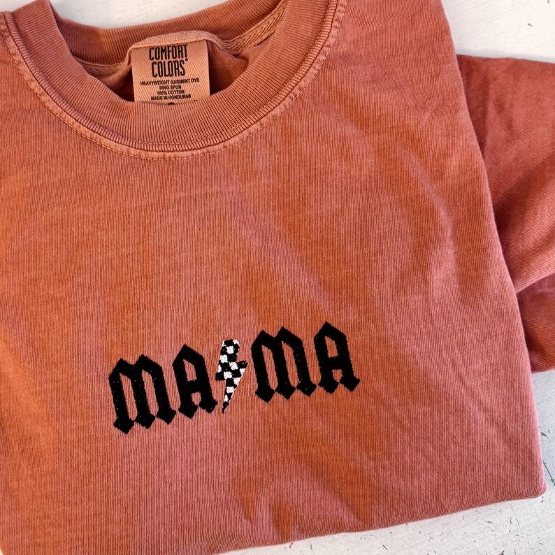 May include: A terracotta-colored Comfort Colors t-shirt with the word "mama" in a gothic font, featuring a checkered lightning bolt graphic. The shirt has a crew neck and short sleeves.