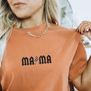 May include: An orange-colored t-shirt with the word "MA*MA" printed in black gothic font. The asterisk is replaced with a black and white checkered flag design. A silver chain necklace is worn around the neck.