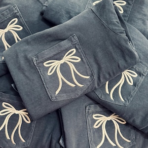 May include: A pile of blue-grey shirts with a pocket detail. Each pocket features an embroidered bow design in a cream color. The shirts are folded, showing the pocket detail prominently. The fabric appears to be a soft, casual material.