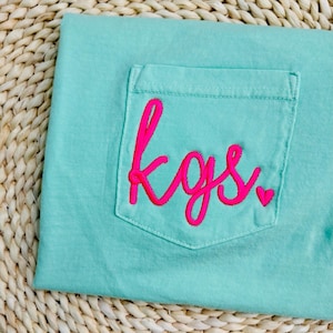 May include: A light turquoise t-shirt with a pocket featuring the embroidered initials "kgs" in bright pink. A small pink heart is embroidered next to the initials. The shirt is made of a soft material.
