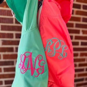 May include: Two hooded jackets, one in a vibrant coral and the other in a light teal. Each jacket features a large, embroidered monogram in a contrasting color. The coral jacket has a teal monogram, while the teal jacket has a pink monogram.
