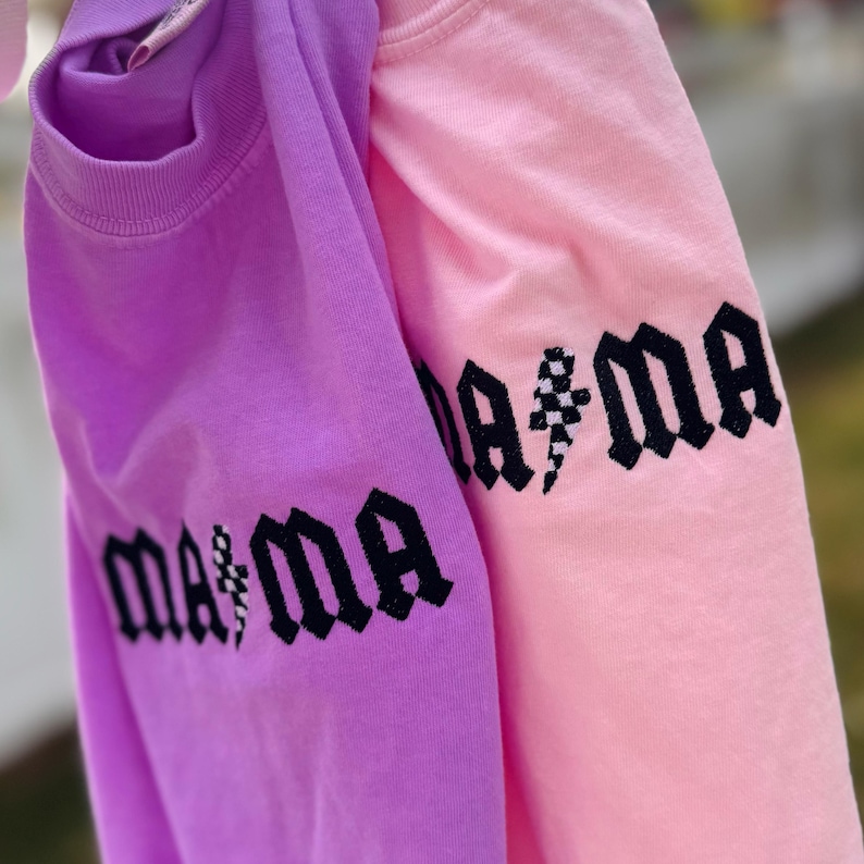 May include: Two sweatshirts, one lavender and one pink, are displayed. The word "mama" is printed in black gothic lettering on the sleeves. A lightning bolt graphic is incorporated into the design. The sweatshirts are made of a soft material.