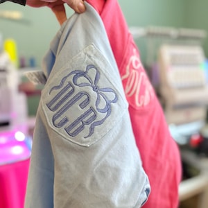 May include: A light blue shirt with a chest pocket featuring an embroidered purple monogram of the letters DCB topped with a bow design. Beside it is a bright pink shirt with a white embroidered monogram of the letters DCB on the chest pocket.