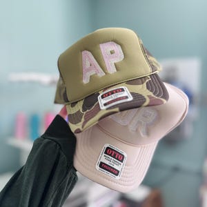 May include: Two trucker hats stacked together. The top hat features a camouflage pattern with a solid olive green front panel and the letters AP in light pink. The bottom hat is solid light pink with the letters JR visible. Both have OTTO labels.
