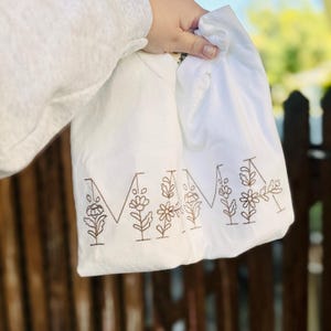 May include: White tote bag with the word "MAMA" embroidered in brown thread. The letters are outlined with small floral designs.