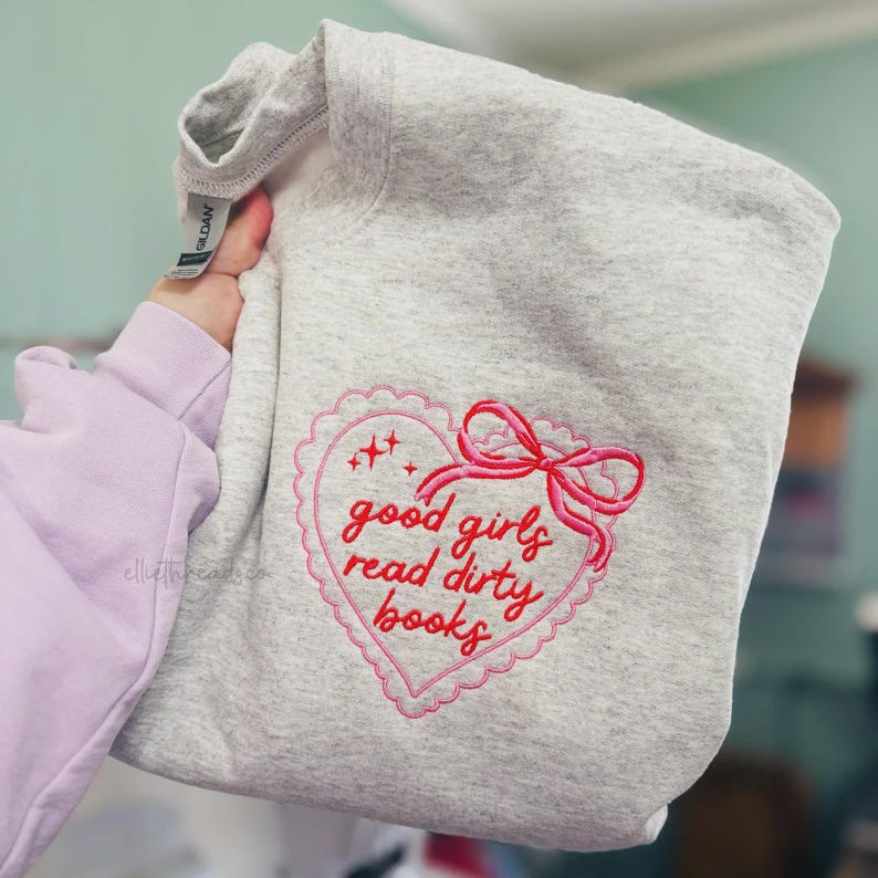 May include: A light gray sweatshirt with a pink embroidered heart design. The heart features a scalloped edge, a bow, and the text "good girls read dirty books" in pink script. The sweatshirt is folded.