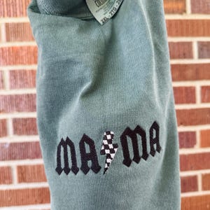 May include: A sage green long-sleeve shirt with the embroidered word "ma ma" in black gothic lettering. A black and white checkered lightning bolt is between the words. The shirt has a tag that says "2XL".
