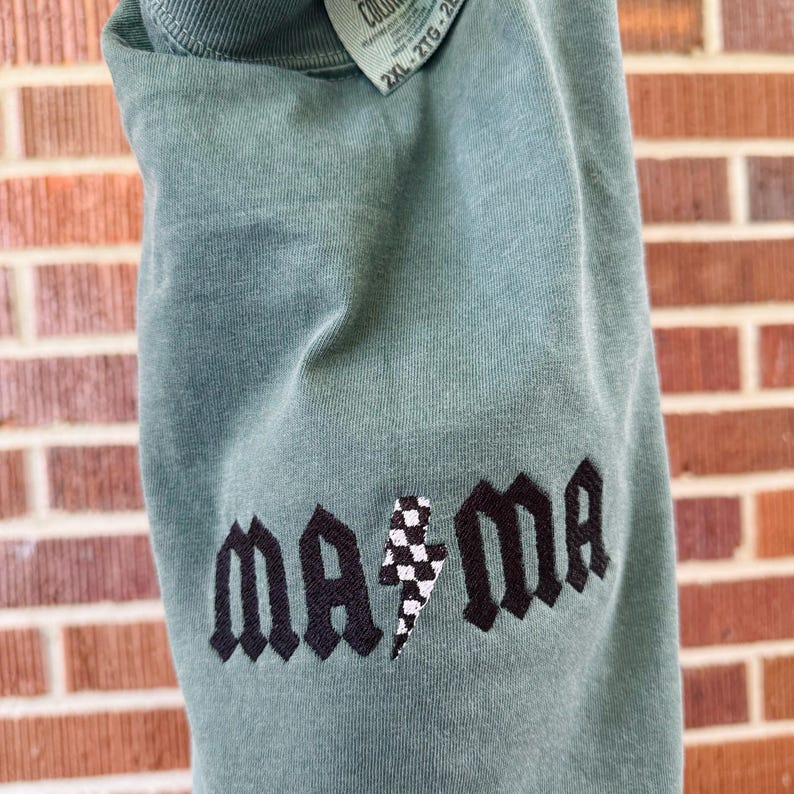 May include: A sage green long-sleeve shirt with the embroidered word "ma ma" in black gothic lettering. A black and white checkered lightning bolt is between the words. The shirt has a tag that says "2XL".