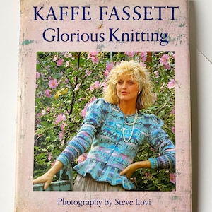 May include: A well-worn book cover for "KAFFE FASSETT Glorious Knitting". The cover features a woman in a colourful knitted jacket, set against a backdrop of pink flowers. The title is in dark blue on a faded pink background.
