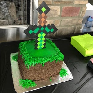 May include: A square-shaped cake decorated with green frosting to look like grass and a brown dirt base. A green and black pixelated sword is stuck into the top of the cake.